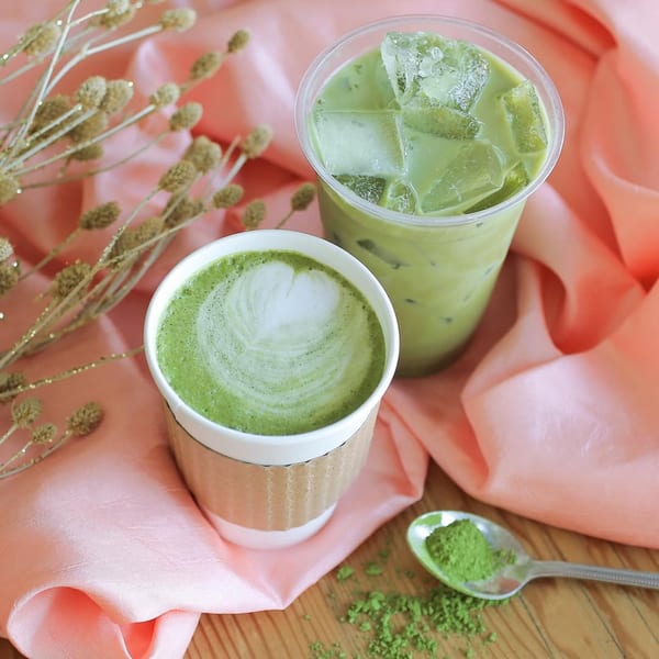 Two matcha drinks, one hot with latte art and one iced, on a wooden table with pink fabric.