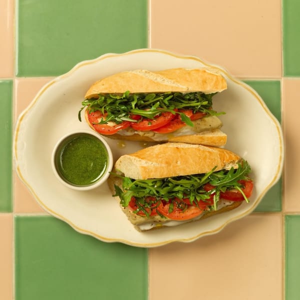 Two baguette sandwiches with arugula, tomatoes, and chicken, served with green sauce on a white plate.