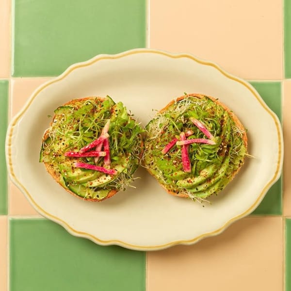 Two avocado toasts with sprouts and radish on a white plate, against green and peach tiles.