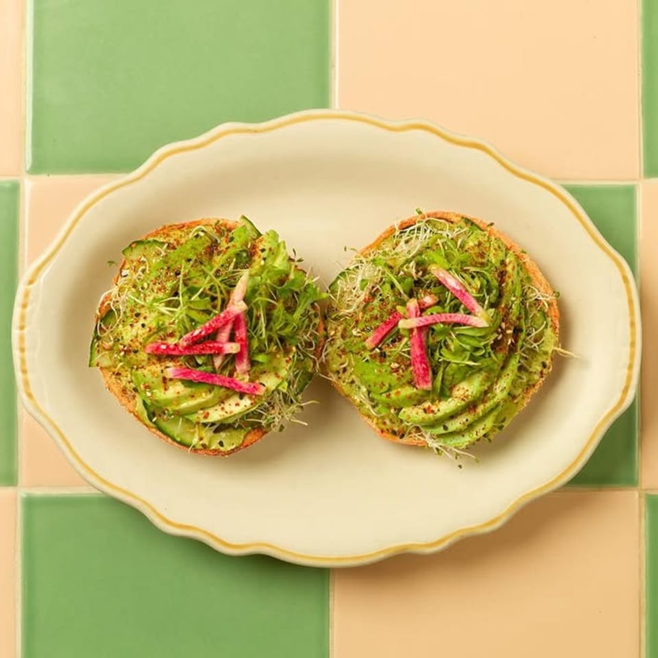 Two avocado toasts with sprouts and radish on a white plate, against green and peach tiles.