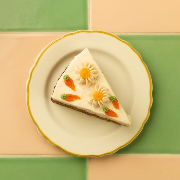 A slice of carrot cake with white frosting, decorated with frosting carrots and daisies, on a white plate.