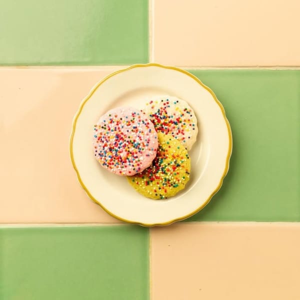 Three colorful, sprinkle-covered cookies on a white plate with a gold rim, against a green and peach tiled background.