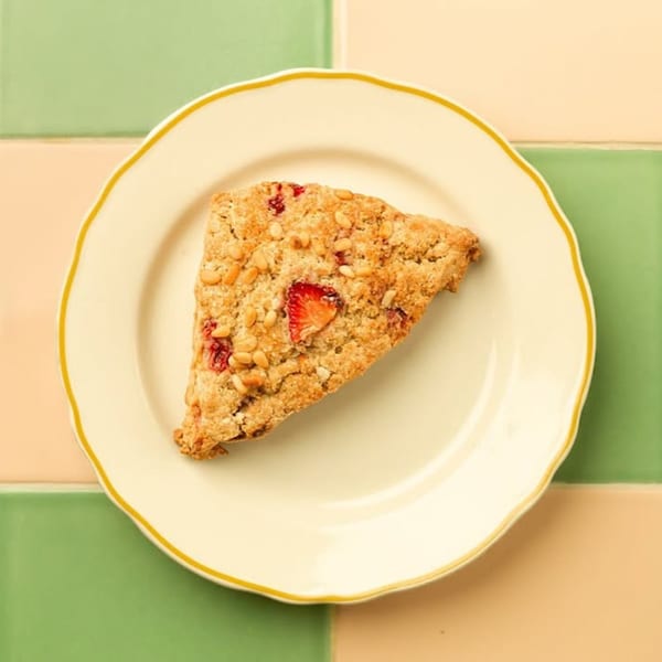 A strawberry scone with pine nuts on a white plate with a yellow rim, against a green and peach tiled background.
